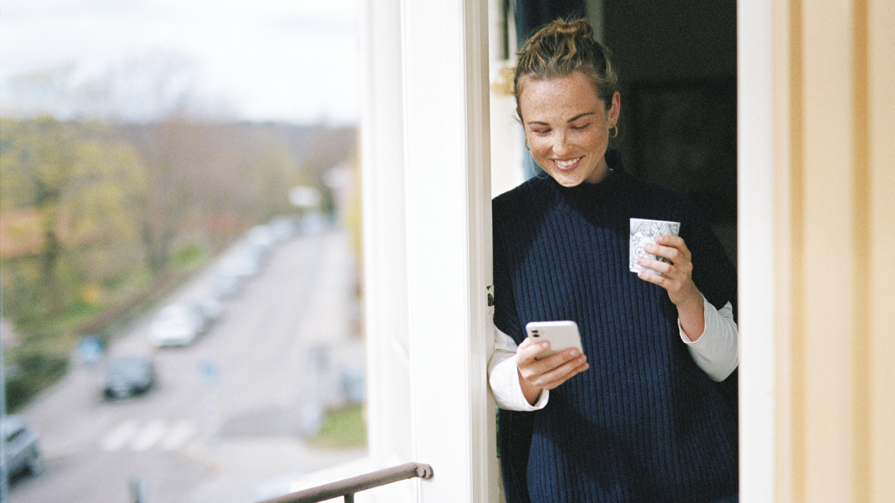 Ung kvinna ler medan hon står på sin balkong med en kaffekopp i handen och tittar på sin mobil