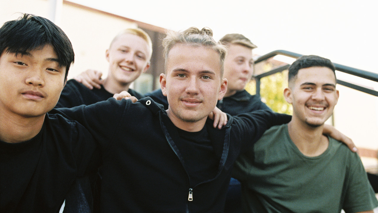 Fem glada unga killar sitter på en trappa vid ett torg 