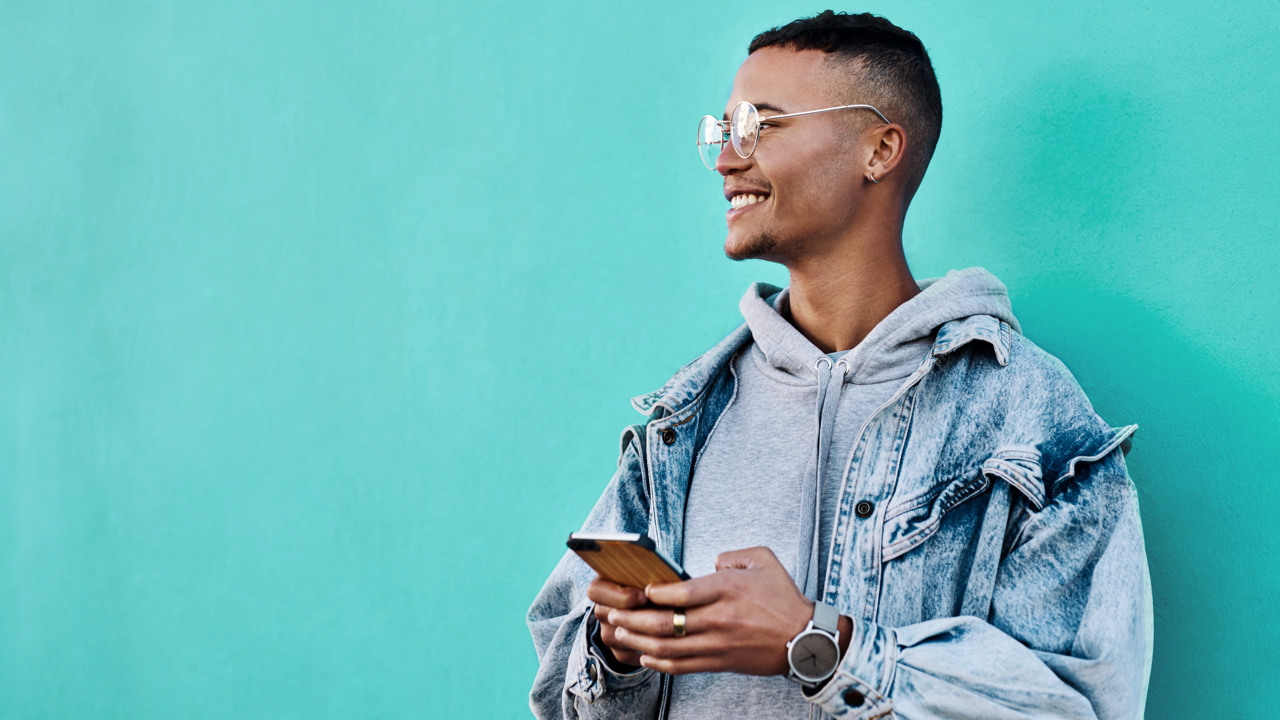 En ung leende man står med sin mobiltelefon mot en turkos vägg.